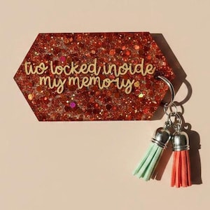 May include: A red glitter keychain with the text "to locked inside my memory". The keychain has a silver ring and two tassels, one mint green and one coral.