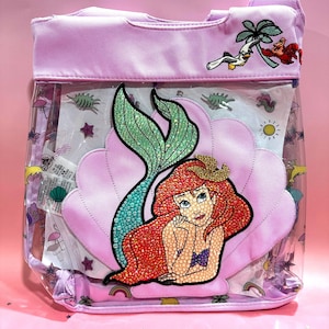 Puede incluir: Una bolsa de pl&aacute;stico transparente con forro de tela morado. La bolsa presenta una gran sirena pelirroja brillante con cola verde, sentada en una concha rosa. La bolsa tambi&eacute;n tiene una peque&ntilde;a imagen bordada de una palmera, una gaviota y un cangrejo.