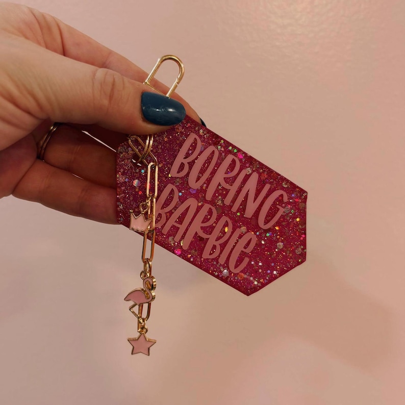 May include: A pink glittery tag keychain with the words "BORING BARBIE" in pink. The keychain has a gold clasp and a gold chain with a pink flamingo, crown, and star charm. The tag is held by a hand with blue nail polish.