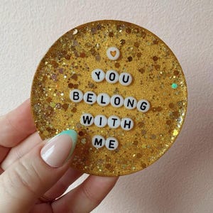 May include: A round, gold glitter coaster with the words "YOU BELONG WITH ME" spelled out in white letter beads. A small heart is above the word "YOU". The coaster is held by a hand with a French manicure.
