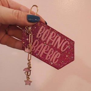 May include: A pink glitter keychain with the words "BORING BARBIE" in a cursive font. The keychain has gold-colored hardware, a small crown charm, a flamingo charm, and a star charm.