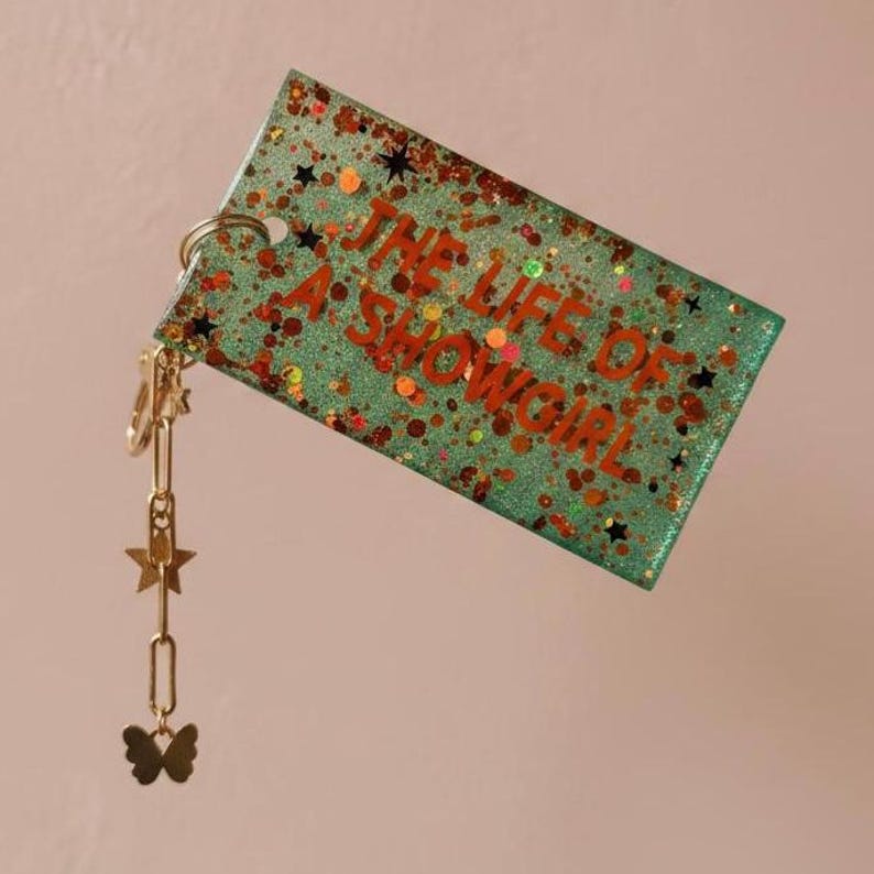 May include: A rectangular keychain with the text "THE LIFE OF A SHOWGIRL" in orange. The keychain is teal with glitter and star accents. A gold chain with star and butterfly charms hangs from the top.