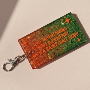 Op de afbeelding: Een rechthoekige sleutelhanger met een glinsterende oranje en groene gradi&euml;nt. De sleutelhanger heeft de tekst "GOT ME DREAMING BOUT A DRIVEWAY WITH A BASKETBALL HOOP" in het oranje. Het heeft een zilveren sluiting.