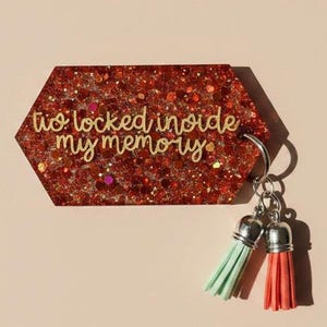 May include: A hexagonal keychain with a red glitter background and the gold text "tio locked inside my memory." It has a silver keyring and two tassels, one mint green and one coral.