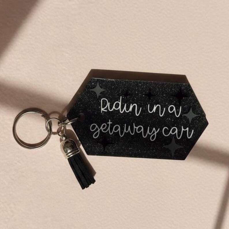 Op de afbeelding: Zwarte zeshoekige sleutelhanger met de tekst "Ridin in a getaway car" in wit script. De sleutelhanger heeft een zilveren sleutelring en een zwarte kwast. De achtergrond is lichtroze.