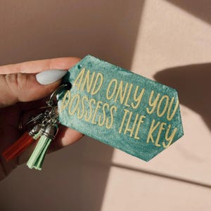 Op de afbeelding: Een turquoise sleutelhanger met de gouden tekst "AND ONLY YOU POSSESS THE KEY". De sleutelhanger heeft rode en groene kwastjes en is bevestigd aan een zilveren sleutelring.