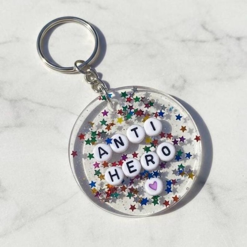 Puede incluir: Un llavero redondo y transparente con las palabras "ANTI HERO" escritas con cuentas de letras blancas. El llavero est&aacute; lleno de confeti en forma de estrella de colores y tiene un llavero plateado.