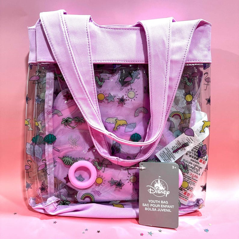 Puede incluir: Una bolsa de pl&aacute;stico transparente con una parte superior de lona morada y un estampado rosa y blanco de varias im&aacute;genes con tem&aacute;tica veraniega. La bolsa tiene dos asas y una etiqueta que dice "Disney Youth Bag Sac Pour Enfant Bolsa Juvenil 3+"