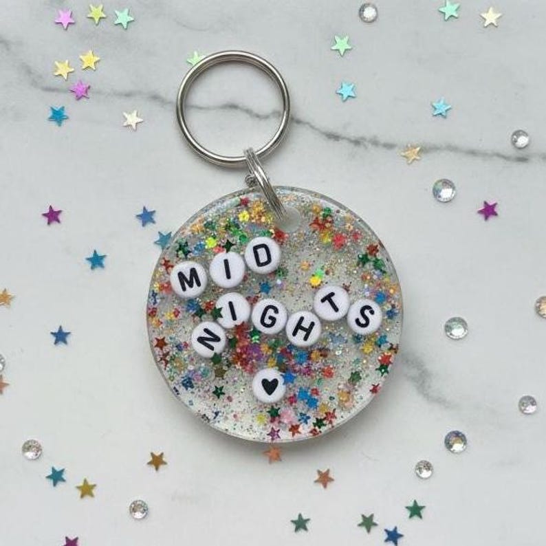 Puede incluir: Un llavero redondo con la palabra "MIDNIGHTS" escrita con cuentas de letras blancas. El llavero est&aacute; lleno de purpurina y confeti colorido en forma de estrella. Un llavero plateado est&aacute; adjunto en la parte superior.