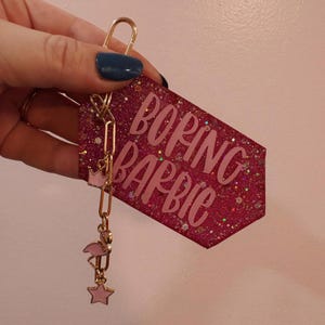 May include: A pink glittery keychain with the words "BORING BARBIE" in light pink. The keychain has a gold-colored chain with a pink crown, flamingo, and star charm. The keychain is held by a person.
