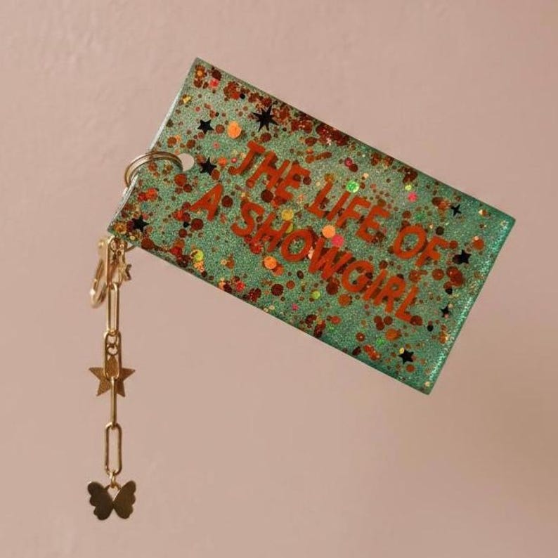 Op de afbeelding: Een rechthoekige sleutelhanger in teal met de tekst "THE LIFE OF A SHOWGIRL" in oranje. De sleutelhanger heeft glitter- en steraccenten. Een gouden ketting met ster- en vlinderbedels hangt aan de sleutelhanger.