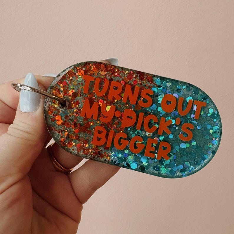 Op de afbeelding: Ovale sleutelhanger met de tekst "TURNS OUT MY PICK'S BIGGER" in oranje. De sleutelhanger is verdeeld in twee secties, &eacute;&eacute;n met oranje glitter en de andere met blauwe glitter. Een zilveren sleutelring is bevestigd.