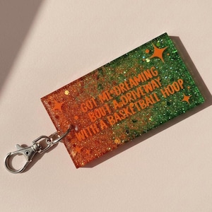 May include: A rectangular keychain with a green and orange glitter design. The keychain has the text "GOT ME DREAMING BOUT A DRIVEWAY WITH A BASKETBALL HOOP" in orange. A silver-colored clasp is attached.