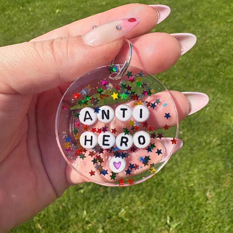 Puede incluir: Llavero redondo y transparente con las palabras "ANTI HERO" escritas con cuentas de letras blancas. El llavero est&aacute; lleno de purpurina colorida en forma de estrella y un peque&ntilde;o coraz&oacute;n morado. Un llavero plateado est&aacute; adjunto.
