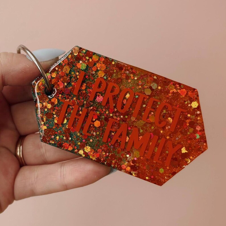 Op de afbeelding: Een glitter sleutelhanger met de tekst "I PROTECT THE FAMILY" in het rood. De sleutelhanger heeft de vorm van een label met een zilveren sleutelring. De glitter is een mix van rood, oranje en groen.