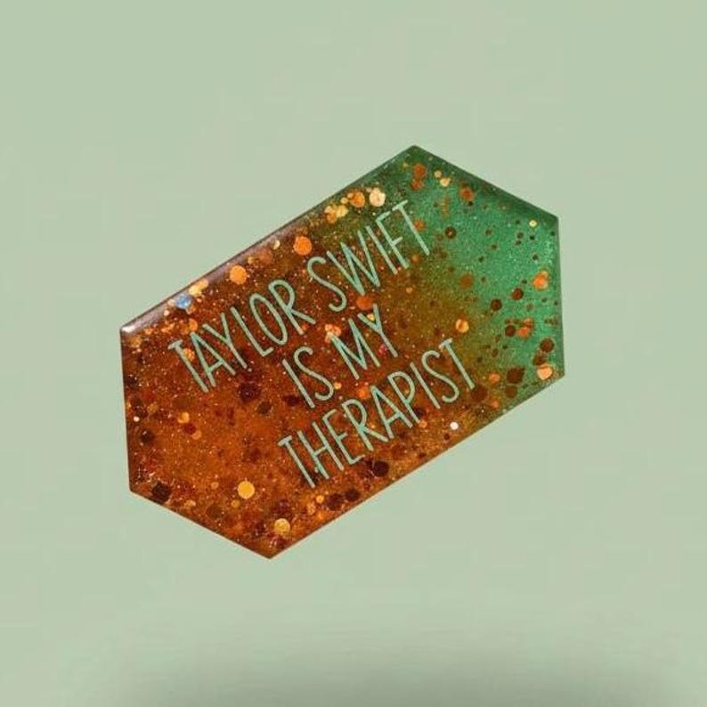 May include: A decorative, geometric-shaped item with the text "TAYLOR SWIFT IS MY THERAPIST." The item has a gradient design, transitioning from a glittery orange to a glittery green. The background is a solid, muted green.