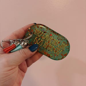 Op de afbeelding: Een turquoise en glitterende sleutelhanger met de tekst "THEY GAVE HER THE KEYS TO THE CITY". De sleutelhanger heeft een zilveren sluiting en twee kwastjes in oranje en turquoise.