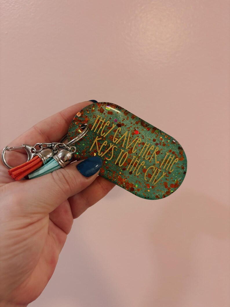 Op de afbeelding: Een turquoise en glitterende sleutelhanger met de tekst "THEY GAVE HER THE KEYS TO THE CITY". De sleutelhanger heeft een zilveren sluiting en twee kwastjes in oranje en turquoise.