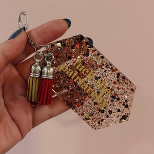Op de afbeelding: Een glitterende sleutelhanger met de gouden tekst "fuck the patriarchy". De sleutelhanger heeft een zilveren ketting en twee kwastjes, &eacute;&eacute;n goud en &eacute;&eacute;n rood.