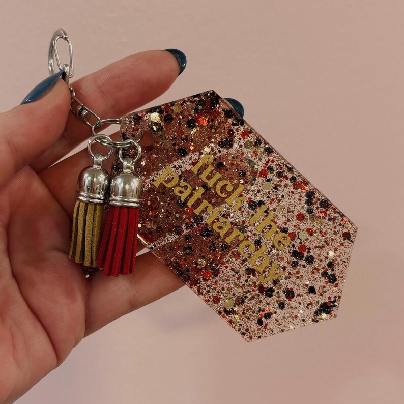 Op de afbeelding: Een glitterende sleutelhanger met de gouden tekst "fuck the patriarchy". De sleutelhanger heeft een zilveren ketting en twee kwastjes, &eacute;&eacute;n goud en &eacute;&eacute;n rood.