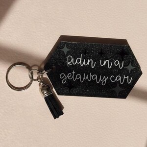 May include: Black hexagonal keychain with the phrase "Ridin in a getaway car" in white script. The keychain has a silver keyring and a black tassel. The background is a light tan color.