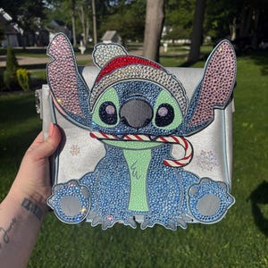 Pu&ograve; includere: Una borsa argentata con un design di personaggio blu scintillante con un cappello da Babbo Natale e un bastoncino di zucchero. Il personaggio ha grandi occhi e una pancia verde. La borsa &egrave; decorata con strass e fiocchi di neve.