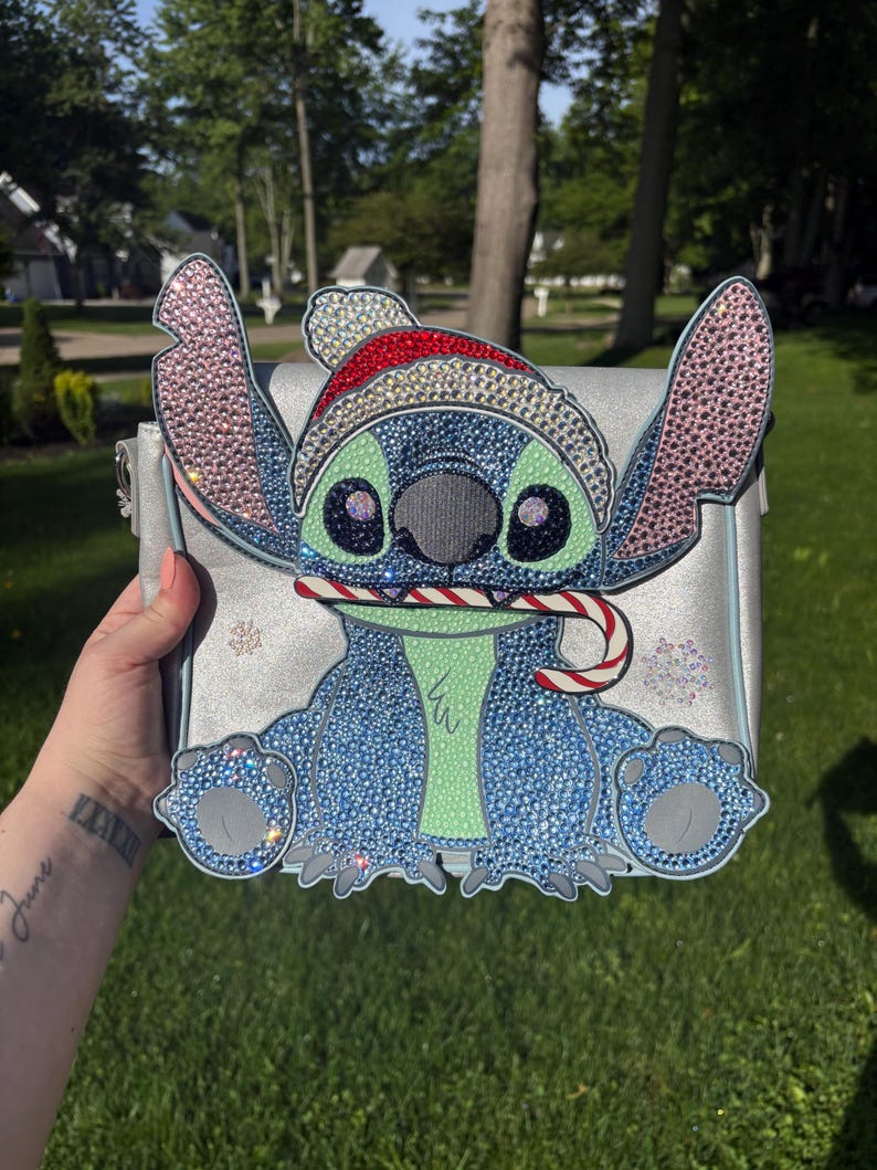 Pu&ograve; includere: Una borsa argentata con un design di personaggio blu scintillante con un cappello da Babbo Natale e un bastoncino di zucchero. Il personaggio ha grandi occhi e una pancia verde. La borsa &egrave; decorata con strass e fiocchi di neve.