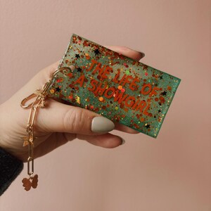 Op de afbeelding: Een rechthoekige sleutelhanger met de tekst "THE LIFE OF A SHOWGIRL" in het rood. De sleutelhanger heeft een groene glitterachtergrond met rode en gouden glitteraccenten en zwarte stervormen. Een gouden ketting met ster- en vlinderbedels is bevestigd.