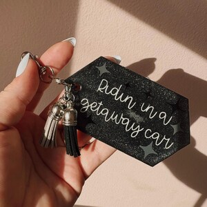 Op de afbeelding: Zwarte glitter sleutelhanger met de tekst "Ridin in a getaway car" in witte letters. De sleutelhanger heeft zilverkleurige hardware en twee kwastjes, &eacute;&eacute;n grijze en &eacute;&eacute;n zwarte. Decoratieve sterren zijn ook afgebeeld.