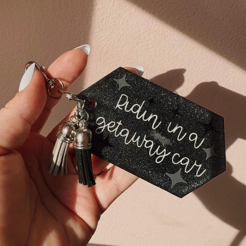 Op de afbeelding: Zwarte glitter sleutelhanger met de tekst "Ridin in a getaway car" in witte letters. De sleutelhanger heeft zilverkleurige hardware en twee kwastjes, &eacute;&eacute;n grijze en &eacute;&eacute;n zwarte. Decoratieve sterren zijn ook afgebeeld.