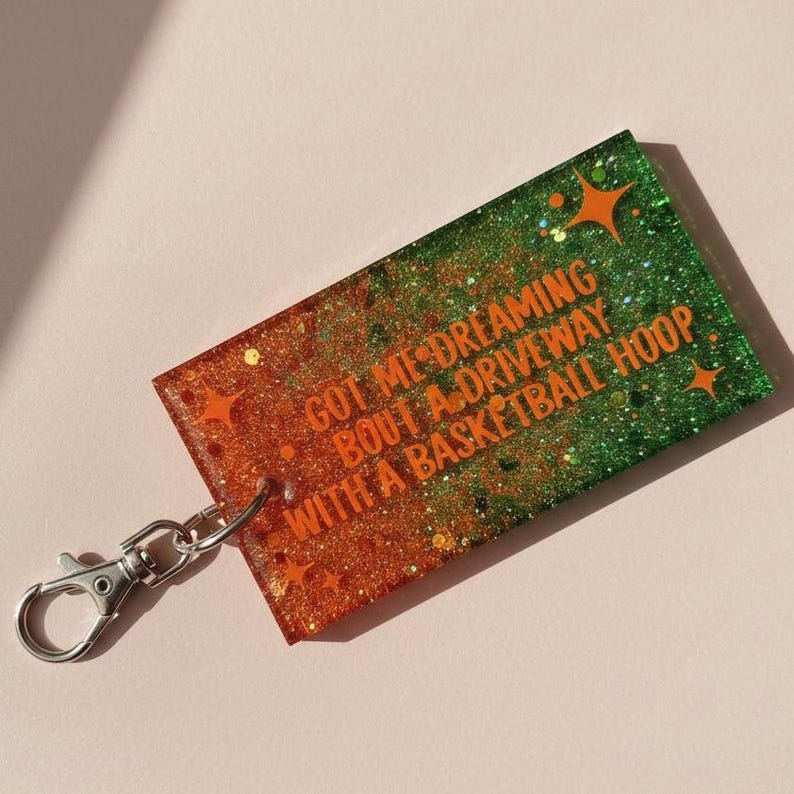 May include: A rectangular keychain with a silver clasp. The keychain is orange and green with glitter and star accents. Text on the keychain reads "GOT ME DREAMING BOUT A DRIVEWAY WITH A BASKETBALL HOOP."