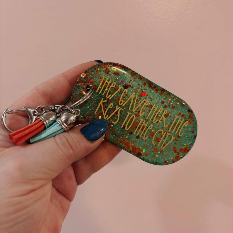 May include: A teal and glitter keychain with the phrase "They Gave Her The Keys To The City" in gold lettering. The keychain has a silver clasp and two tassels, one red and one teal.