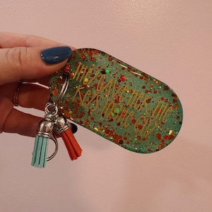 May include: A teal glitter keychain with the text "They Gave Her The Keys To The City" in gold lettering. The keychain has two tassels, one teal and one orange.