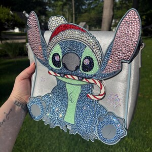 Pu&ograve; includere: Borsa a tracolla argentata con un design Stitch scintillante. Il personaggio blu &egrave; adornato con strass e un cappello da Babbo Natale, che tiene in mano un bastoncino di zucchero. La borsa ha una tracolla argentata ed &egrave; decorata con fiocchi di neve.