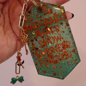 Op de afbeelding: Een teal en glitterende sleutelhanger met de tekst "AND BABY THAT'S SHOW BUSINESS FOR YOU" in het rood. De sleutelhanger heeft goudkleurige hardware en bedels, waaronder een strik, sleutels en een roos.