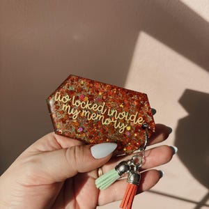 Op de afbeelding: Een glitterende, rood-oranje sleutelhanger met de tekst "to locked inside my memory". De sleutelhanger heeft een zilveren sluiting en twee kwastjes, &eacute;&eacute;n mintgroen en &eacute;&eacute;n koraal.