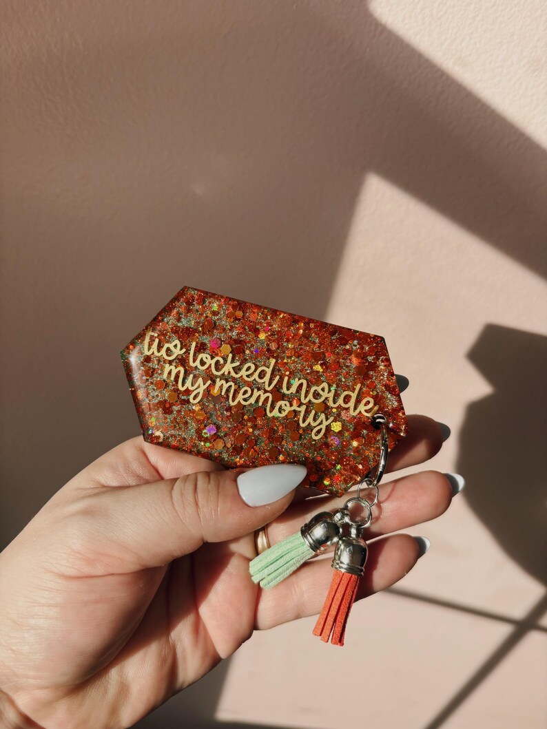 Op de afbeelding: Een glitterende, rood-oranje sleutelhanger met de tekst "to locked inside my memory". De sleutelhanger heeft een zilveren sluiting en twee kwastjes, &eacute;&eacute;n mintgroen en &eacute;&eacute;n koraal.