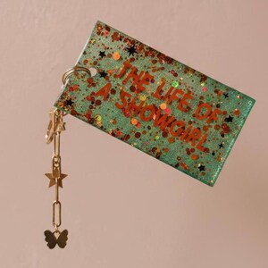 May include: A rectangular keychain with the text "THE LIFE OF A SHOWGIRL" in orange lettering. The keychain is teal with glitter and star accents. A gold-colored chain with star and butterfly charms is attached.