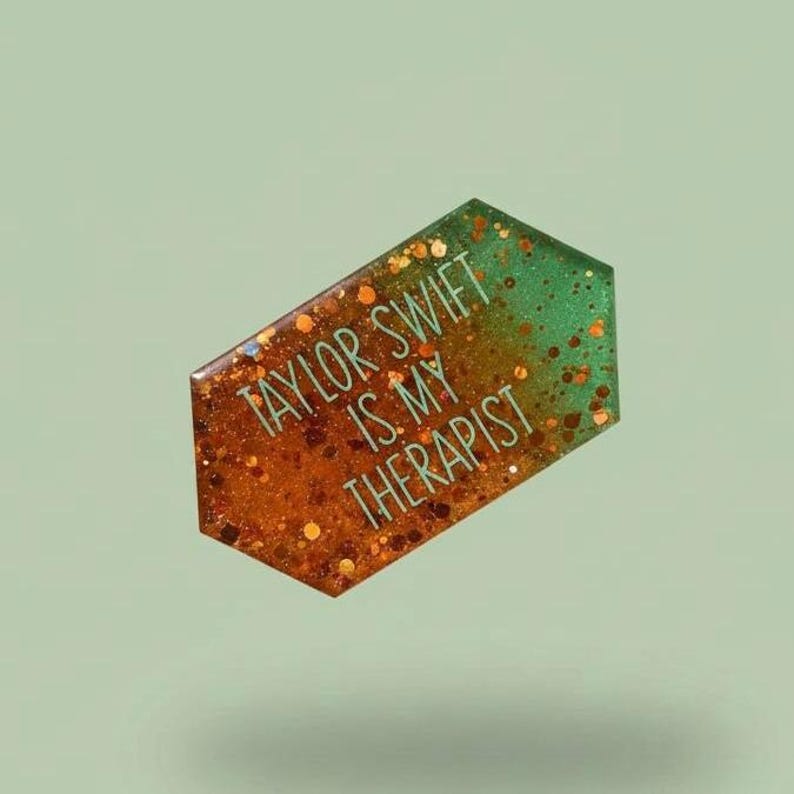 May include: A glittery, geometric-shaped badge with the text "TAYLOR SWIFT IS MY THERAPIST" in teal. The badge has a gradient of orange to green with gold glitter accents, set against a soft green background.