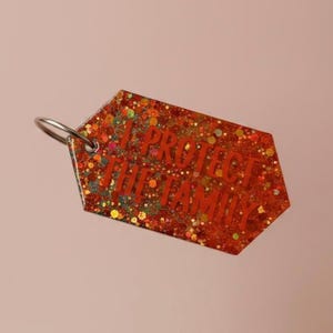 Op de afbeelding: Een oranje glitter sleutelhanger met de rode tekst "I PROTECT THE FAMILY". De sleutelhanger heeft een zilveren metalen ring en een geometrische vorm. De glitter bevat groene, gele en oranje spikkels.