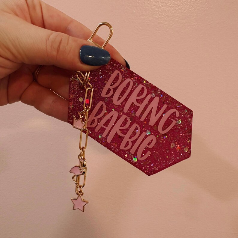 May include: A pink glittery keychain with the words "BORING BARBIE" in a cursive font. The keychain has a gold clasp and a chain with a pink crown, flamingo, and star charm.