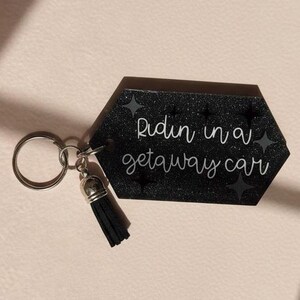 May include: Black glitter keychain with the phrase "Ridin in a getaway car" in white script. The keychain has a silver key ring and a black tassel.