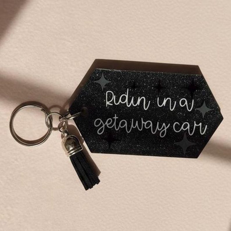 May include: Black glitter keychain with the phrase "Ridin in a getaway car" in white script. The keychain has a silver key ring and a black tassel.