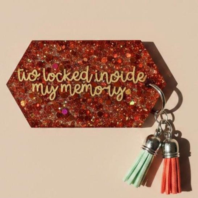 Op de afbeelding: Een rode glitter sleutelhanger met de tekst "to locked inside my memory". De sleutelhanger heeft een zilveren ring en twee kwastjes, &eacute;&eacute;n mintgroen en &eacute;&eacute;n koraal.