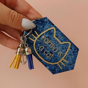 Op de afbeelding: Een blauwe glitter sleutelhanger met de gouden tekst "Karma is a cat". Het katvormige ontwerp heeft gele en blauwe kwastjes.