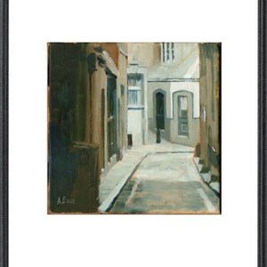 May include: Framed artwork depicting a narrow European street scene. The painting features buildings in shades of brown, gray, and white, with a cobblestone street leading into the distance. The artwork is in a black frame.