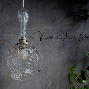 May include: A clear glass pendant light fixture with a silver chain and a textured glass shade. The light is hanging from a ceiling against a grey wall.