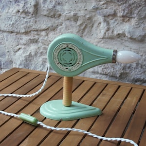 Puede incluir: Un secador de pelo vintage Handy Hannah verde y blanco con un cable blanco y una bombilla en el extremo. El secador está sobre una mesa de madera.