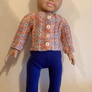 May include: A doll with blonde hair wearing a long-sleeved, multi-colored knit cardigan with four white buttons, solid blue pants, and white sneakers with purple stripes. The cardigan features a textured cable knit pattern in shades of orange, blue, and white.