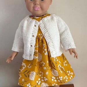May include: A doll with blonde hair wearing a mustard yellow dress featuring a white floral pattern, a white knitted cardigan with four buttons, and white sneakers with purple stripes. The doll has blue eyes and pink lips.